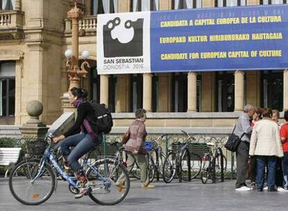 Varios ciudadanos pasan ante un cartel sobre la candidatura donostiarra instalado en el Ayuntamiento de San Sebastián.