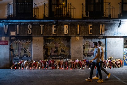 Dos personas pasan frente a las flores en homenaje a Conchi, asesinada en su tienda de la plaza de Tirso de Molina.