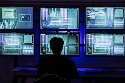 Hacker con el rostro cubierto en su sala de operaciones con múltiples pantallas y cables.