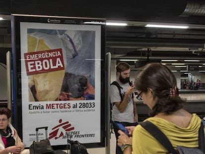 Unos turistas esperan un tren en la estación de Barcelona Sants junto a un cartel de lucha contra el ébola en 2014.