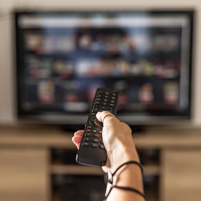 Close up of a person holding a control remote with a television screen on the background. A woman holding a remote control switches programs on the TV in the background.