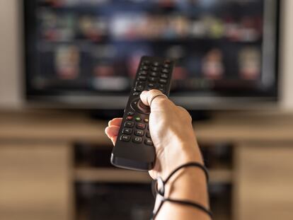 Close up of a person holding a control remote with a television screen on the background. A woman holding a remote control switches programs on the TV in the background.