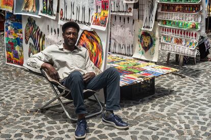 Peter, artista congoleño residente en Sudáfrica desde hace más de dos décadas, posa en su silla de camping, junto a sus pinturas, en la plaza de Green Market, en Ciudad del Cabo. Pincha en la imagen para ver la fotogalería completa.