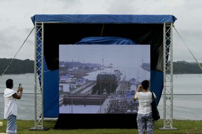 Dos espectadores observan en una pantalla instalada en las inmediaciones del Canal la entrada del carguero.
