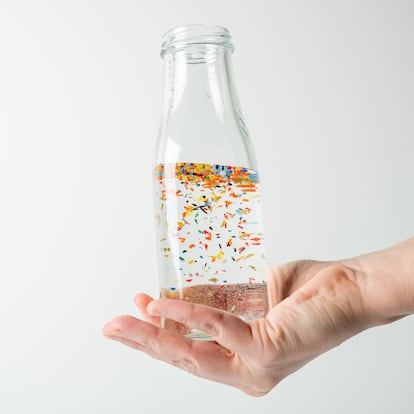 Close-up of hand gripping transparent glass bottle filled with water, where tiny microplastic particles are visibly floating, emphasizing pollution concerns