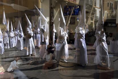 Encapuchados de la hermandad de La Candelaria se reflejan en el cristal de un restaurante durante la procesión por las calles de Sevilla, el 16 de abril de 2019.