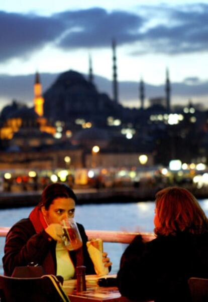 Dos jóvenes en una terraza cerca del puente de Galata, con la silueta de la mezquita de Sulemaniye al fondo.