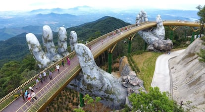 Suspendido sobre dos manos gigantes, el puente de Cau Vang (puente de oro) emerge como un enorme lazo de seda dorada entre los bosques de bambú de Ba Na Hills, una estación balneario construida en 1919, durante la etapa colonial francesa, en las montañas cerca de Da Nang, en el centro de Vietnam. La estructura, una pasarela de madera de 150 metros flanqueada por relucientes barandillas doradas (del estudio vietnamita TA Landscape Architecture), fue inaugurada el pasado junio y ya se ha convertido en uno de los lugares más fotografiados del país asiático. Es la última atracción del gran complejo turístico Sun World, donde hay réplicas de castillos y jardines franceses y un teleférico de casi seis kilómetros de largo.