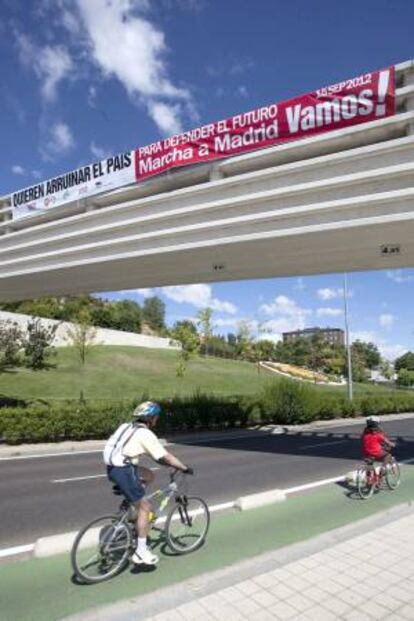 Dos ciclistas pasan por debajo de la pancarta colocada por los sindicatos CC.OO, UGT, CSI-F, USO y STECyL-Intersindical en las proximidades del Museo de la Ciencia de Valladolid, mediante la que promocionan la marcha del 15 de septiembre a Madrid, donde se manifestarán e instarán al Gobierno a que promueva un referéndum para las medidas económicas y reformas que está adoptando.