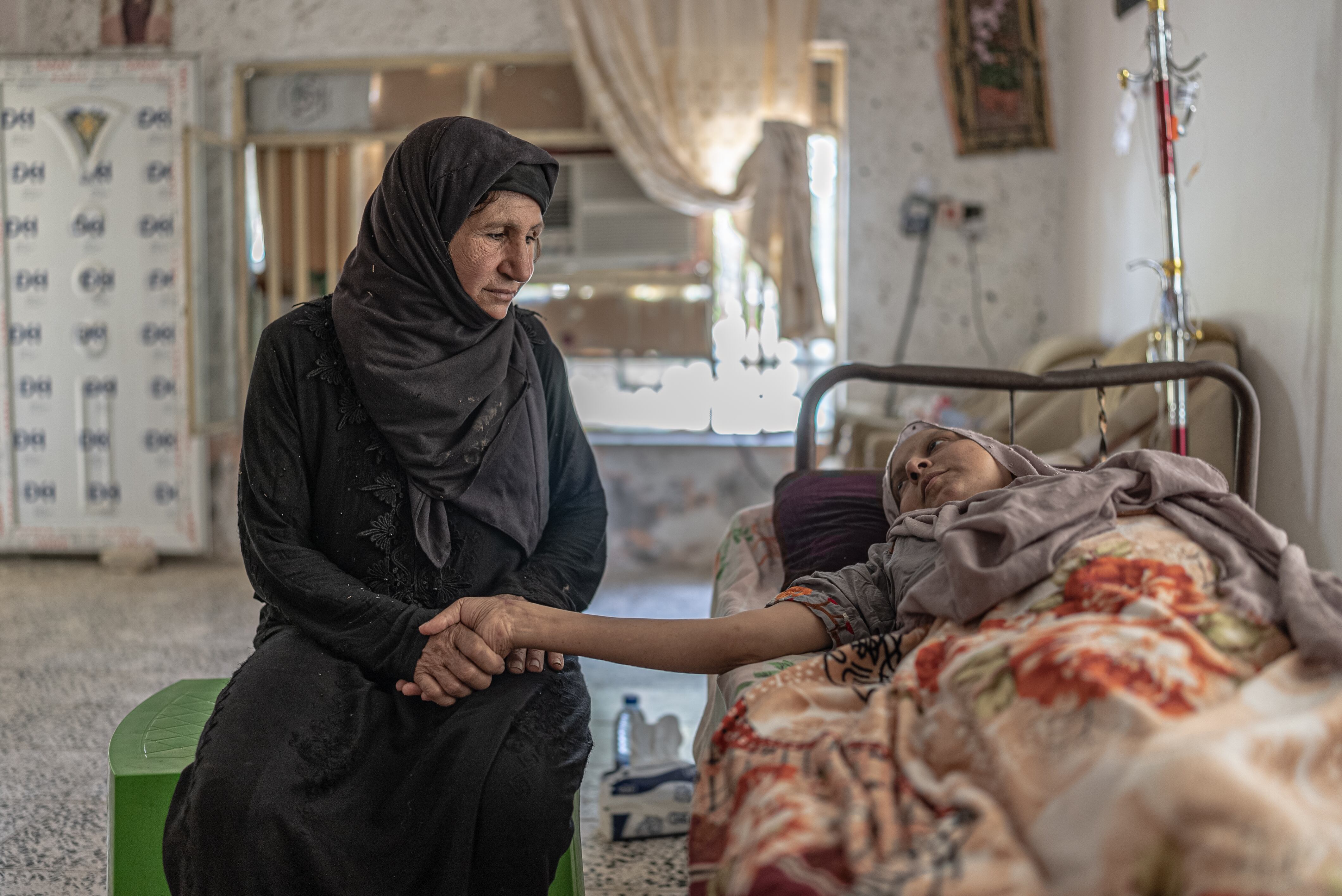 Fátima sostiene la mano de su hija Dika, de 27 años. Dos de sus hijas fueron alcanzadas por metralla durante un ataque del Daesh y se encuentran en casa, ya que la familia no tiene recursos para costear un ingreso en algún hospital. Pincha en la imagen para ver la fotogalería completa. 
