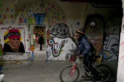 Un hombre se mueve en bicicleta por un pasillo de La Tabacalera, con las paredes decoradas con pinturas y ante una habitación donde se ve a una mujer. El centro cuenta con un taller de arreglo de bicicletas y es común ver este tipo de vehículos apartados en su interior.