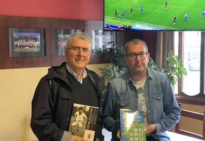 Xosé María Lema y su sobrino Xosé María Rei Lema muestran sus libros sobre el fútbol de la Costa, en un bar de Baio, con fotografías del equipo local de fondo y el pasado Alavés-Celta de LaLiga Santander en la televisión.