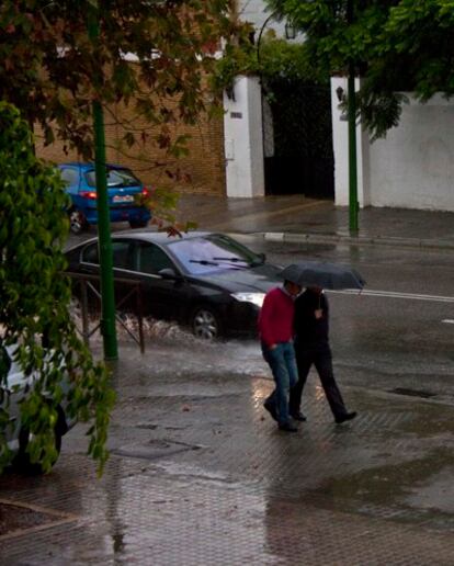 Peatones de Sevilla pasean mientras un coche cruza un charco ocasionado por las fuertes lluvias.