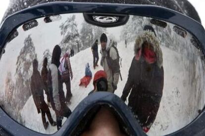 A través del reflejo de unas gafas de ventisca se ve a los visitantes jugar con sus hijos y deslizarse en trineo en el puerto de Navacerrada.