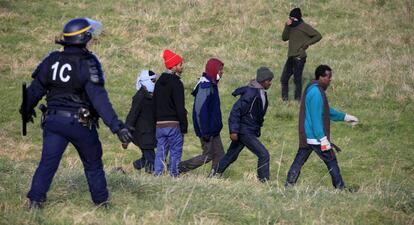Un polic&iacute;a franc&eacute;s antidisturbios vigila este jueves a un grupo de migrantes en un campo cerca de la ciudad de Calais.