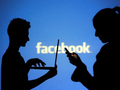 Dos personas, una con un ordenador y otra con un móvil, frente a una pantalla con el logo de Facebook.