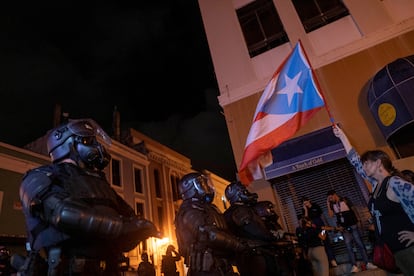 En un comunicado, la Policía de Puerto Rico informó este viernes que cinco agentes resultaron heridos y cuatro personas fueron detenidas durante las protestas. En la imagen, una manifestante sostiene una bandera puertorriqueña frente a un grupo de policías antidisturbios.