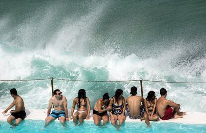 Varias personas permanecen sentadas en el borde de la piscina Icebergs junto al mar mientras una ola se eleva gracias al viento en la playa de Bondi, en Sídney (Australia).