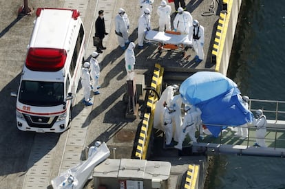 Los infectados han sido trasladados a hospitales en tierra, mientras que el resto de pasajeros y tripulación deberán permanecer aislados en el barco y ser sometidos a nuevas pruebas. En la imagen, los oficiales con un traje de protección escoltan a una persona (debajo de la sábana azul) que estaba a bordo del crucero 'Diamond Princess' y que dio positivo por coronavirus.