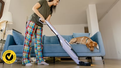 Una mujer pasando la aspiradora mientras su perro mira desde el sofá.