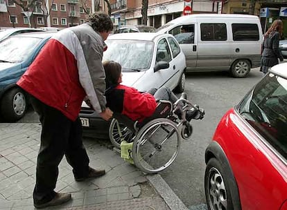 Una discapacitada y su acompañante avanzan entre coches mal aparcados en Madrid.