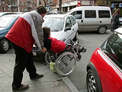 Una discapacitada y su acompañante avanzan entre coches mal aparcados en Madrid.