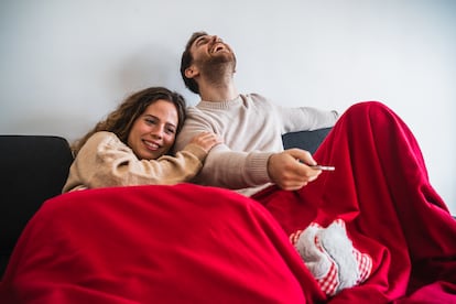 Una pareja, tapada con una manta, ve la televisión desde el sofá.