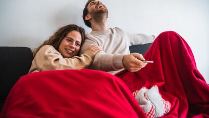 Una pareja, tapada con una manta, ve la televisión desde el sofá.