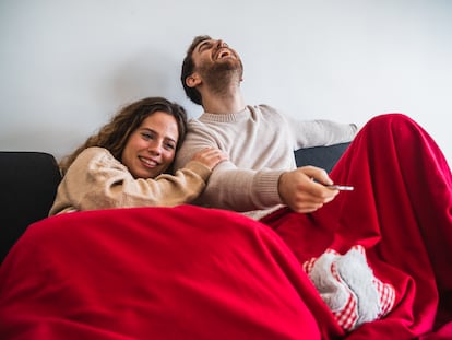 Una pareja, tapada con una manta, ve la televisión desde el sofá.