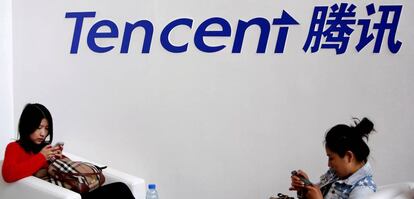 Dos mujeres juegan en su telefono móvil bajo un cartel de la empresa Tencent.