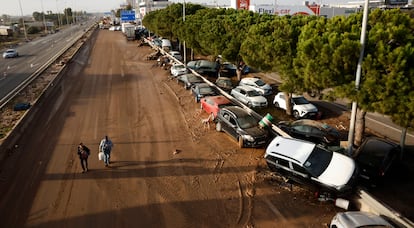 Dos personas caminan por la V-31 en Sedaví (Valencia) junto a decenas de coches apilados contra el quitamiedos, este jueves.