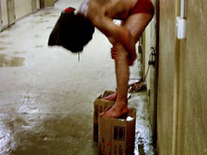 Un reo, con una capucha en la cabeza y un tobillo encadenado a una puerta, es obligado por sus captores a mantener el equilibrio sobre dos cajas de cartón en un pasillo de la prisión de Abu Ghraib.