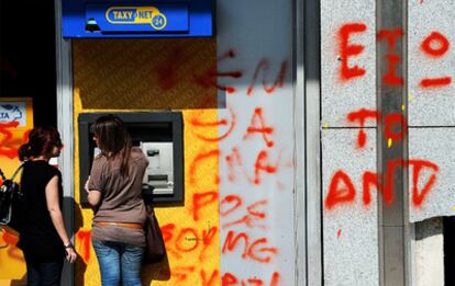 Unas jóvenes griegas sacan dinero en un cajero en el que han escrito <i>Fuera FMI</i>. El país vive hoy nuevas protestas contra el programa de austeridad.