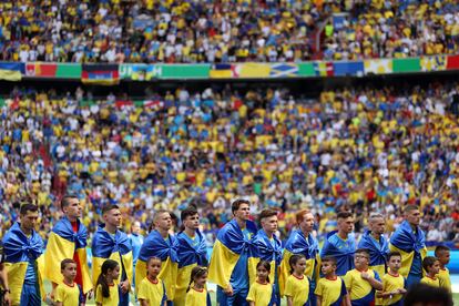 Jugadores ucranios escuchan el himno de su país, envueltos en su bandera.