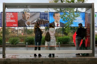 Vecinos de Sevilla esperan un autobús frente a dos carteles de los partidos mayoritarios en una mañana de lluvia.