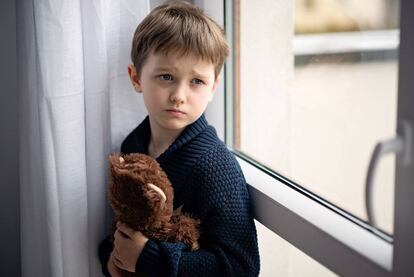 Niño está abrazando a su osito de peluche. De pie junto a la ventana.