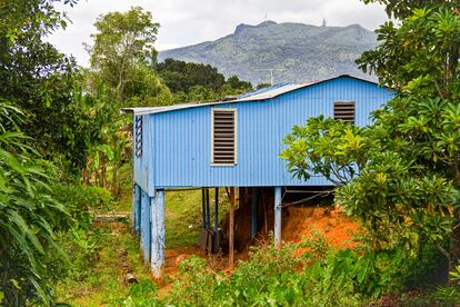 La imagen 'Casita Azul (antes del huracán María)', tomada en Canóvanas, Puerto Rico, en 2013. La casa de madera azul rodeada de árboles y plantas se asienta sobre pilotes.