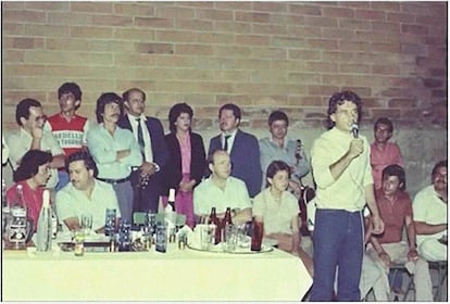 Carlos Lehder hablando, durante un evento en donde al fondo de la fotografía se puede observar a Pablo Escobar.