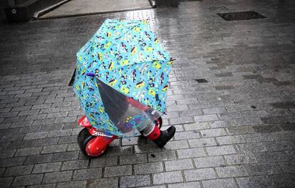 Un ni&ntilde;o se protege de la lluvia bajo un paraguas mientras disfruta con su moto de juguete en una calle de Pamplona.