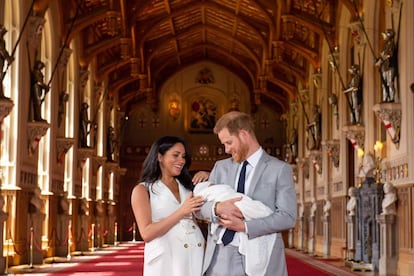 El príncipe mira embelesado a su pequeño, mientras Meghan Markle sonríe y le coloca el gorro blanco, a juego con la toquilla.