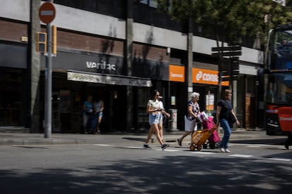 Cuatro personas cruzan la avenida Mare de Déu de Montserrat, frente a dos grandes supermercados que antiguamente fueron una sucursal de La Caixa. 