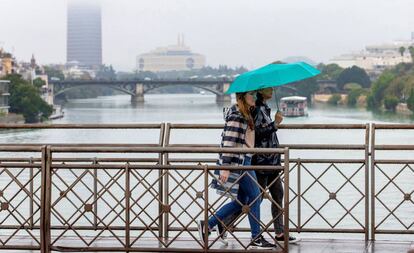 Dos personas atraviesan el puente de San Telmo, con el puente de Triana y Torre Sevilla al fondo, en un desapacible domingo de lluvia en Sevilla.