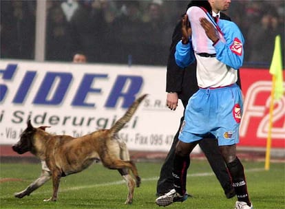 El jugador del Catania Mark Esudei se protege el rostro del humo de las bengalas mientras un perro del sistema de seguridad cruza ante él.