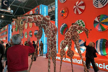 Las dos jirafas que promocionan en Fitur 2003 el futuro parque natural Senda Viva, de Navarra, están animadas por actores de la compañía Xirriquiteula. Al fondo, el colorido espacio de la Comunidad Valenciana.