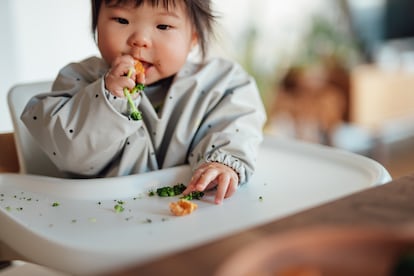 Una niña come trozos de brócoli y de salmón sentada en una trona.