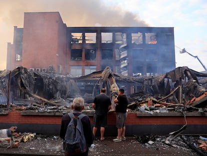 Tres ciudadanos miran este viernes los restos de un edificio en Roubaix, de la empresa Tessi, que fue quemado la tercera noche de disturbios en Francia originados tras la muerte de un menor de 17 años a manos de la policía.