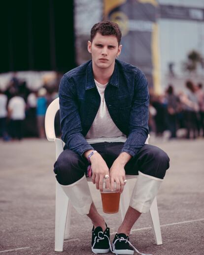 Joan espera sentado a su grupo con chaqueta vaquera de Levi's, camiseta de Bershka, jeans con dobladillo de Loewe y bisutería vintage. Las zapas con suela de goma y cordones son de Victoria. 