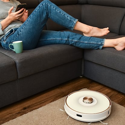 Una mujer viendo el móvil mientras el robot aspiradora limpia la casa.
