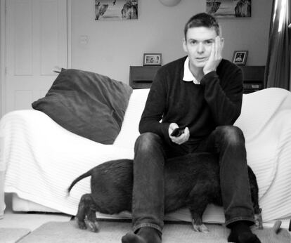 El novelista Matt Whyman en su habitación con uno de sus cerdos pasando por debajo de sus piernas.