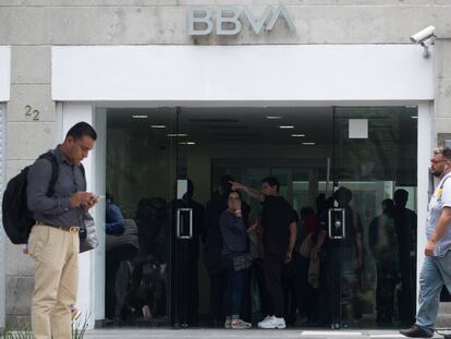 Usuarios frente a una sucursal de BBVA en Ciudad de México que revisan su celular y hacen operaciones bancarias.